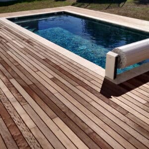 Terrasse en bois moderne bordant une piscine rectangulaire aux eaux bleues, avec volet roulant partiellement enroulé.